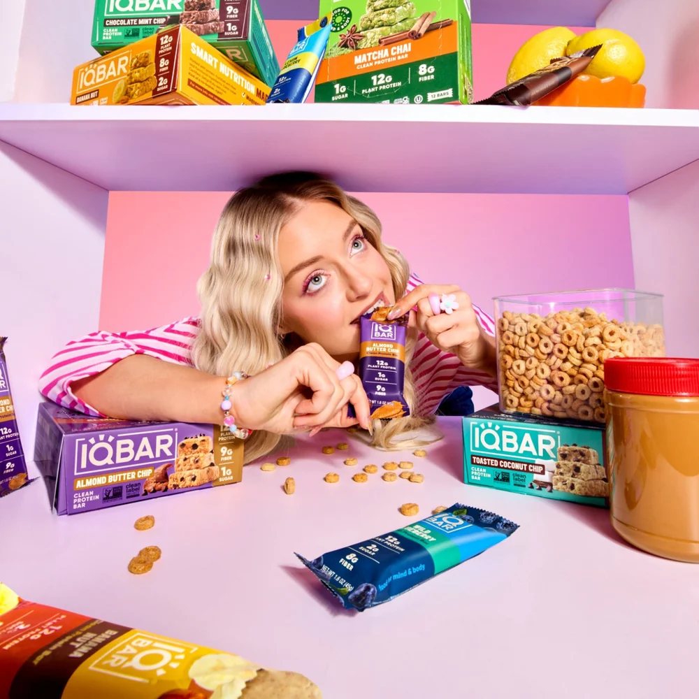 Woman biting an IQBAR Almond Butter Chip bar in a pantry stocked with IQBAR boxes